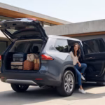 Dark gray Toyota Grand Highlander Hybrid parked in a modern driveway with the rear hatch open showing luggage while a couple opens the rear doors.
