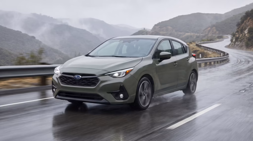 A green 2026 Subaru Impreza hatchback driving confidently along a wet, curving mountain highway under overcast skies.