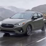 A green 2026 Subaru Impreza hatchback driving confidently along a wet, curving mountain highway under overcast skies.