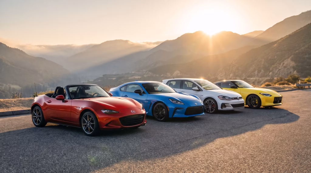 A red Mazda MX-5 Miata, blue Toyota GR86, white Volkswagen Golf GTI, and yellow Nissan Z parked together on a scenic mountain road at sunrise.