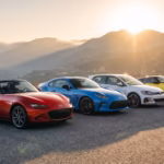 A red Mazda MX-5 Miata, blue Toyota GR86, white Volkswagen Golf GTI, and yellow Nissan Z parked together on a scenic mountain road at sunrise.